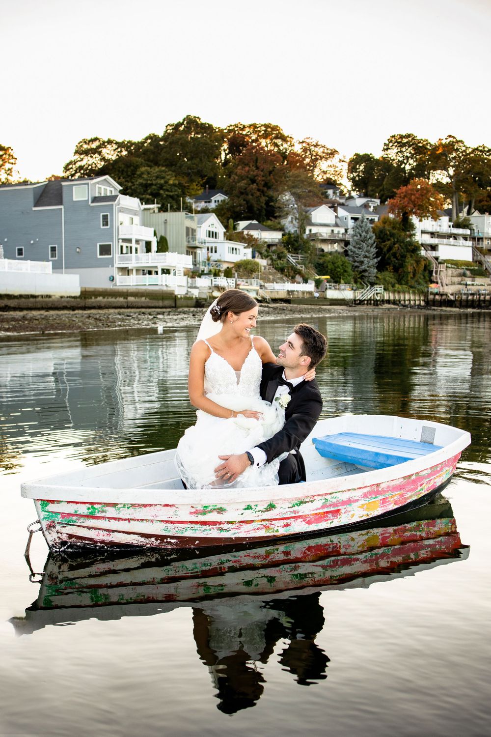 A couple in wedding attire sits in a small boat on calm waters at sunset near coastal homes.