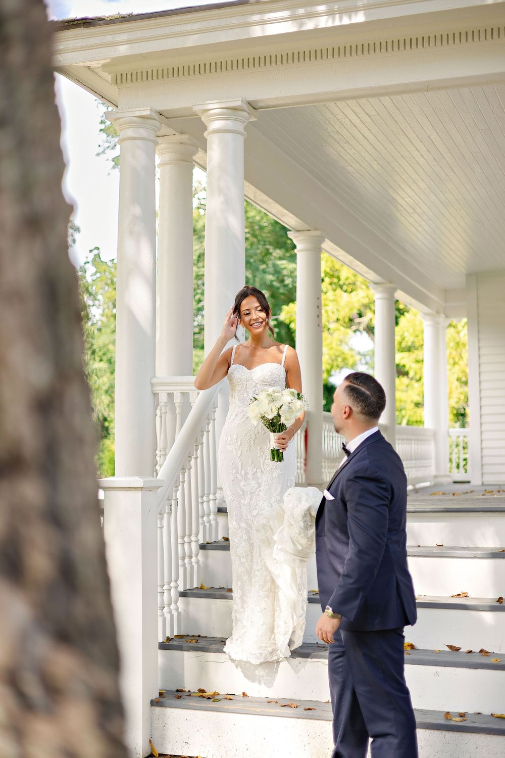 Bride in white lace gown holding bouquet on porch steps, groom in navy suit smiling up at her.