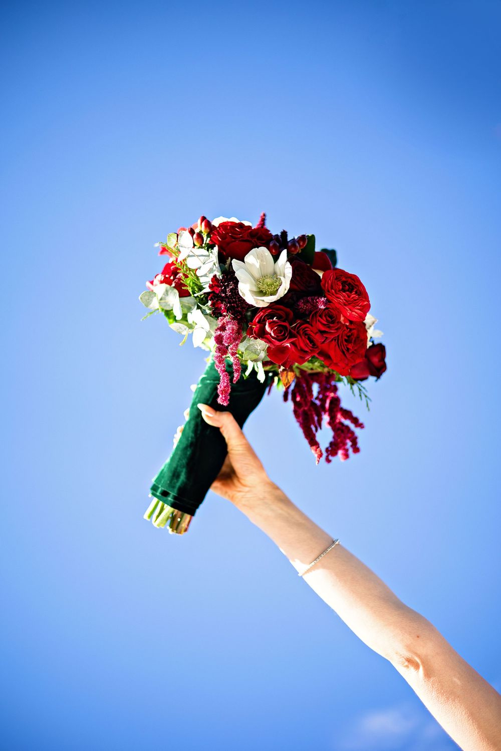 A hand holds up a vibrant bridal bouquet with red and white flowers against a bright blue sky.