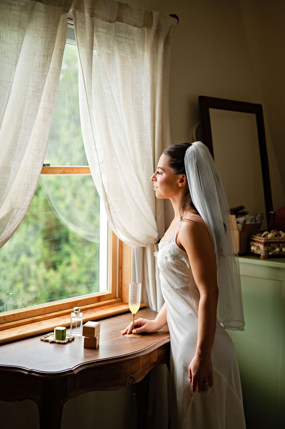Bride in white gown and veil gazes out window beside antique table with champagne glass on her wedding day.
