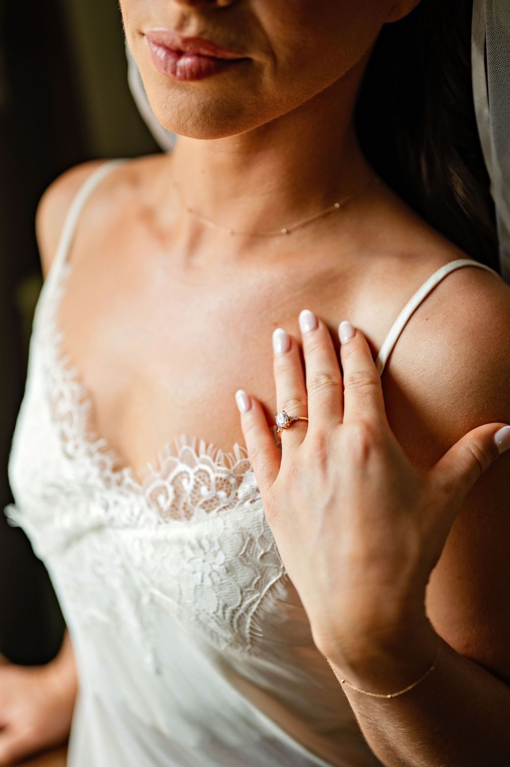 Bride in white lace wedding dress showcasing elegant diamond engagement ring with delicate gold bracelet and necklace.