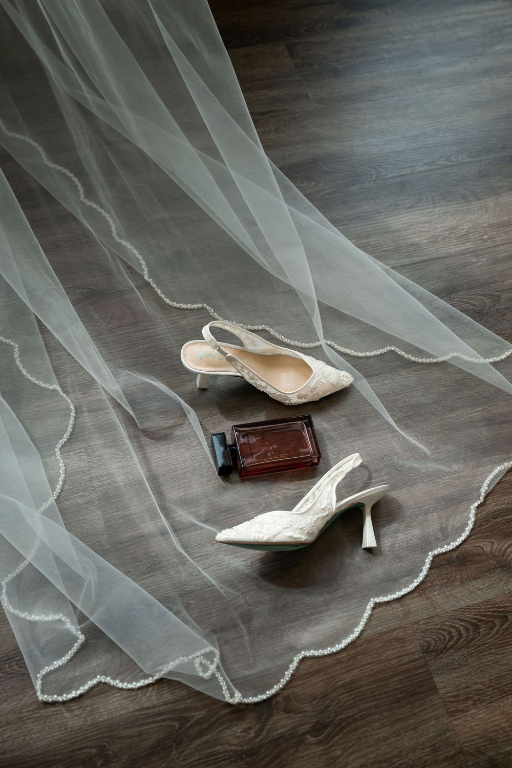 Bridal accessories flat lay featuring white slingback heels, perfume bottle, and sheer veil with scalloped edges on wood floor.