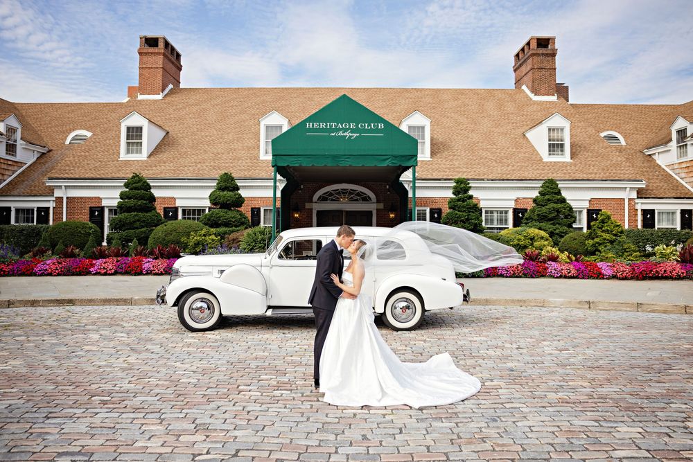 Bride and groom kissing in front of a vintage white car at the Greystone Club wedding venue.