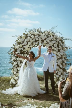 Newlyweds celebrating under luxurious white flower arch with ocean backdrop.