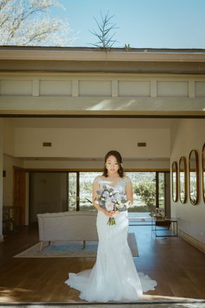 A woman in a white wedding dress holding a bouquet stands in a sunlit room.