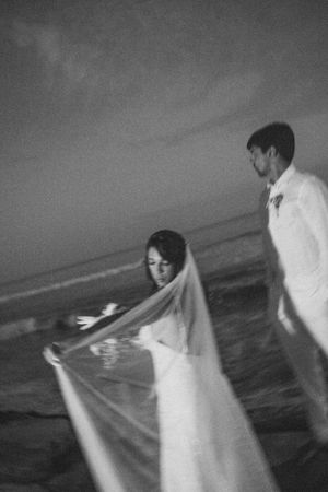 Ethereal black and white scene of couple with flowing wedding dress on beach at dusk.