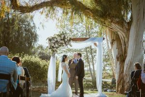 A wedding ceremony under a floral arch with draped fabric and willow trees.