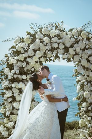 Bride and groom sharing romantic moment under stunning white floral arch by ocean.