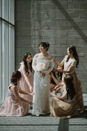 Bridesmaids in pink dresses help adjust the bride's lace gown near a large window with gray brick wall.