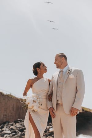 A romantic beach wedding photo capturing a couple in cream-colored attire with seabirds flying overhead.