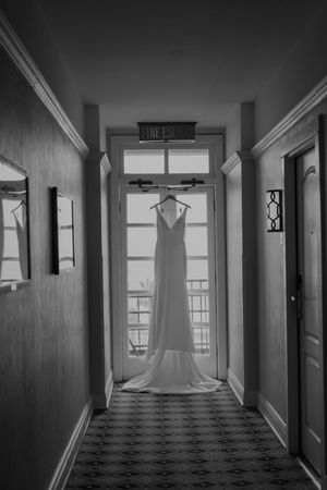 A white wedding dress hangs in silhouette against a bright window at the end of a hallway with patterned carpet.