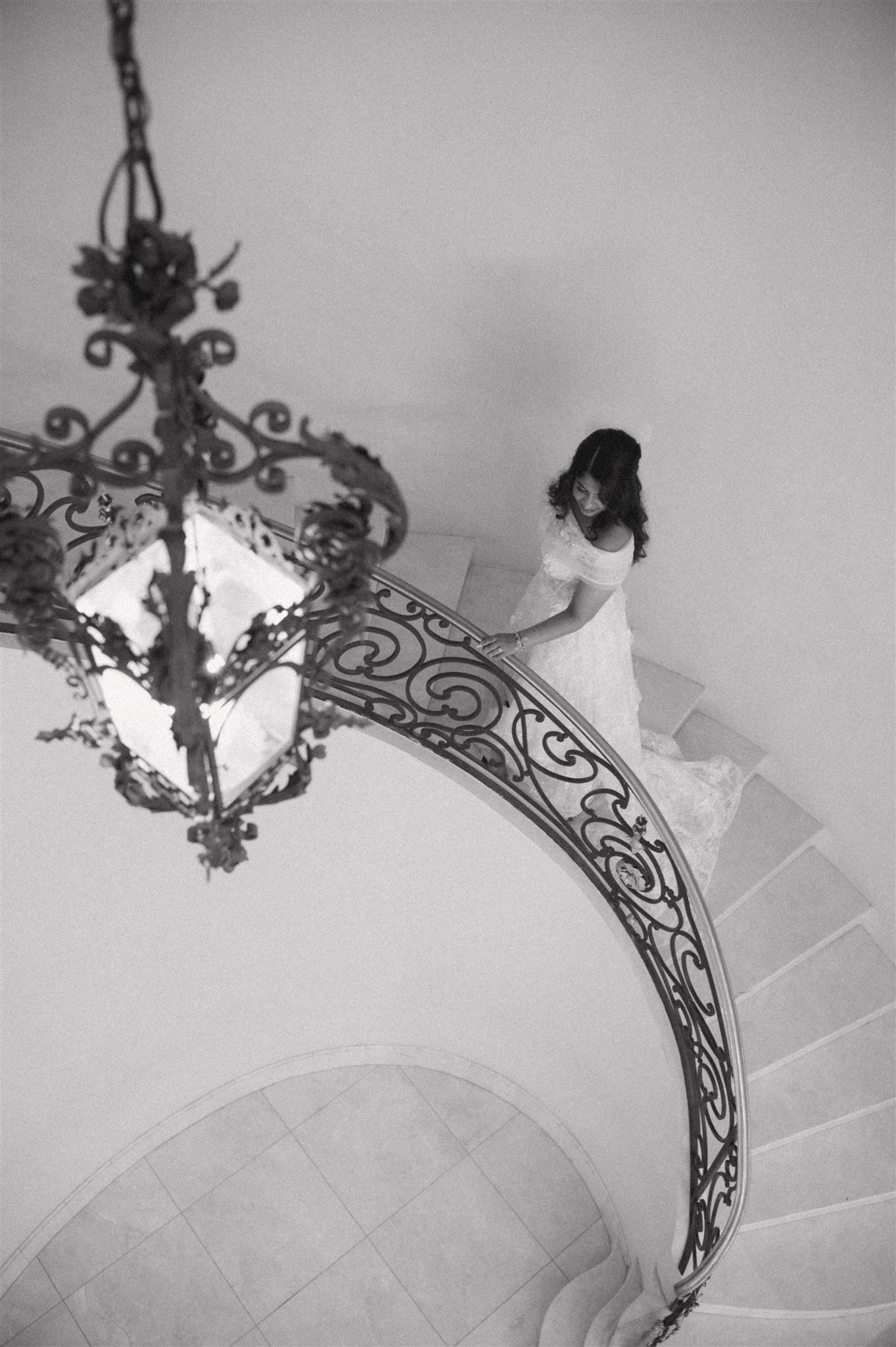 A dramatic black and white photo captures a bride descending a curved staircase beneath an ornate chandelier.