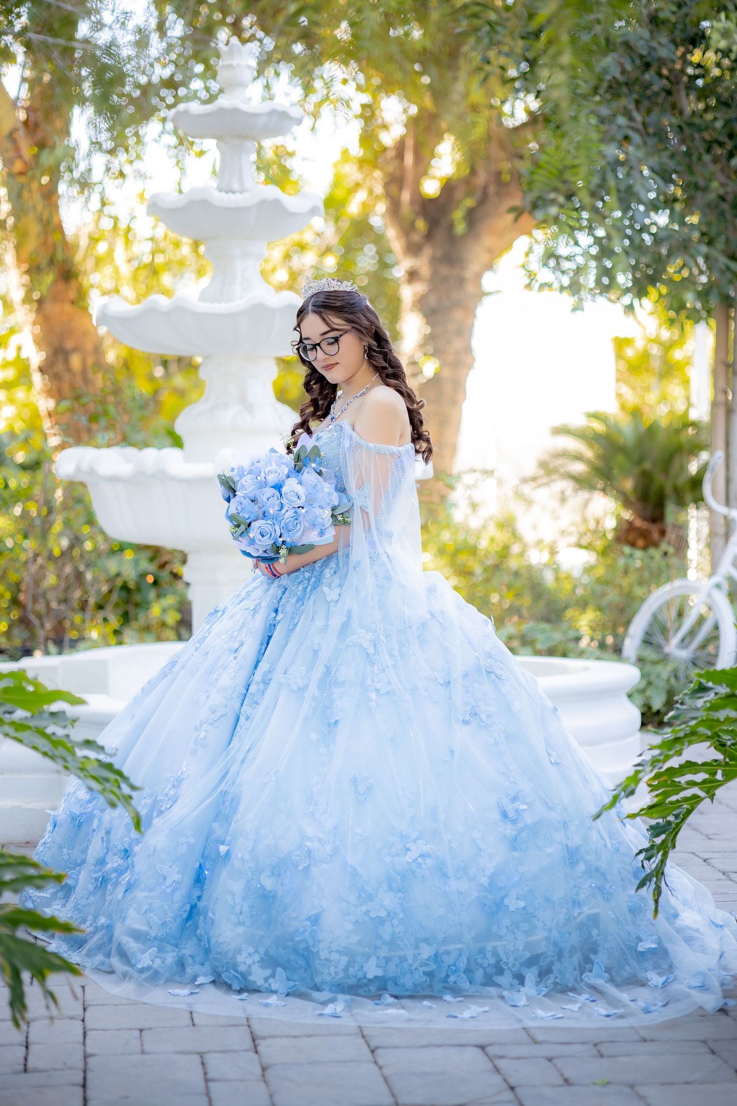 Young woman in light blue quinceañera ball gown holds hydrangea bouquet near a white fountain outdoors.