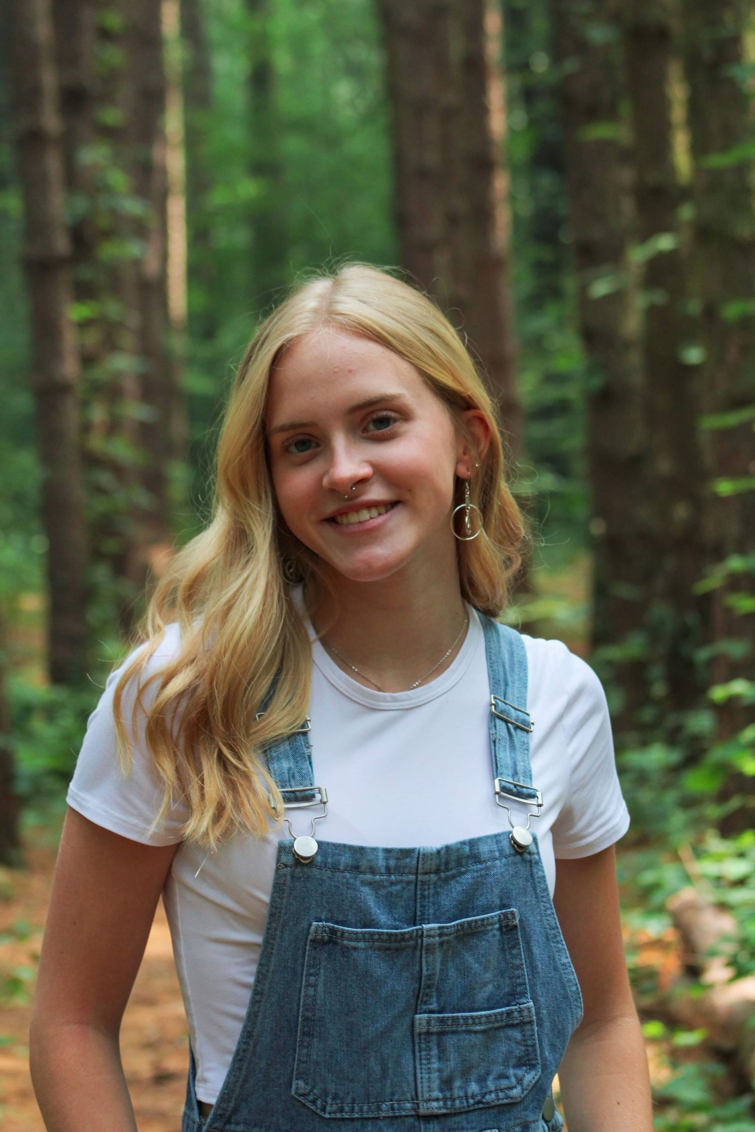 Person in denim overalls and white t-shirt poses in wooded setting.