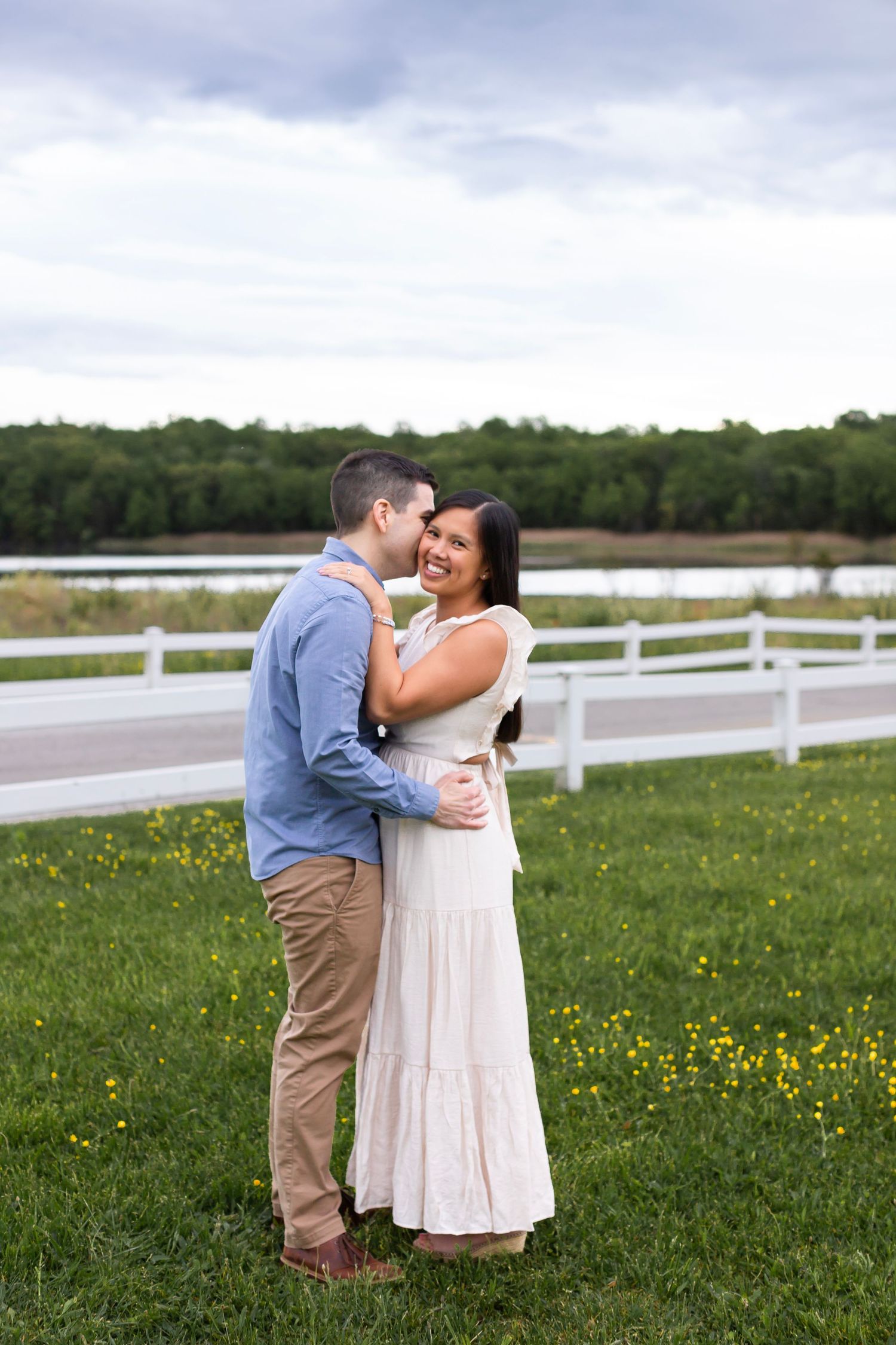 A romantic moment between a couple in front of a white fence and lake on a cloudy day.