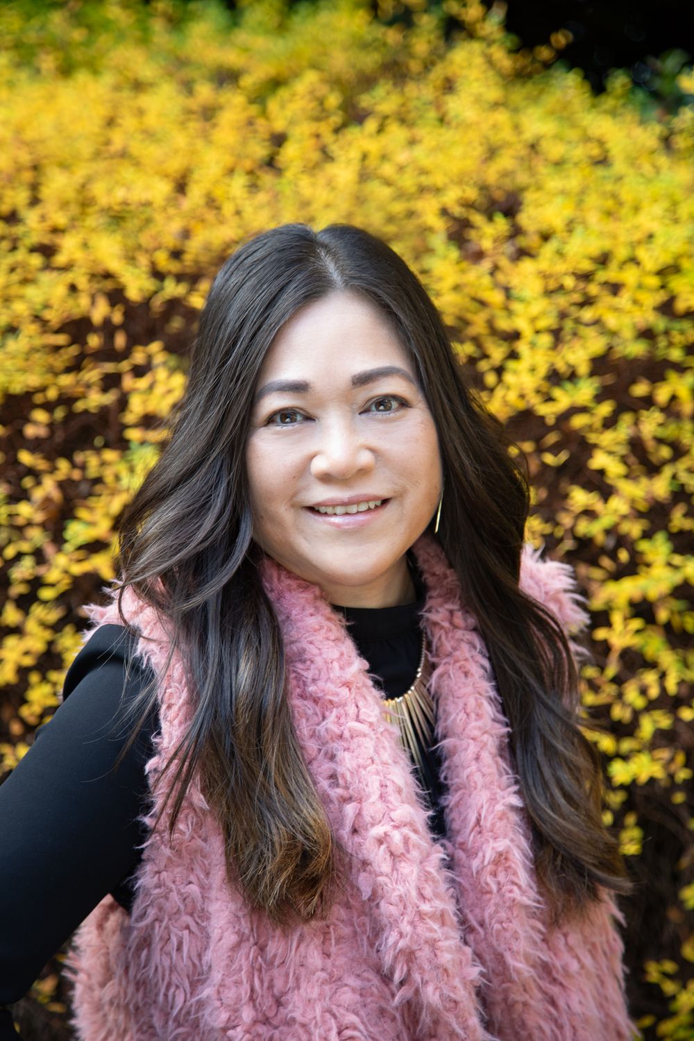 Outdoor headshot portrait of woman in pink vest with black top and a yellow flowering bush background