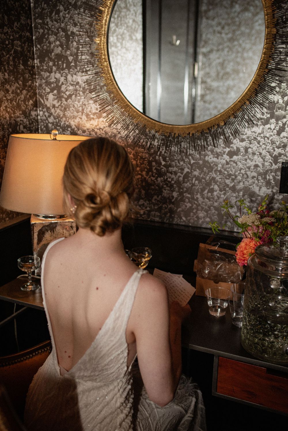 A back view of someone in a white dress sitting at a vanity with a round mirror and table lamp.