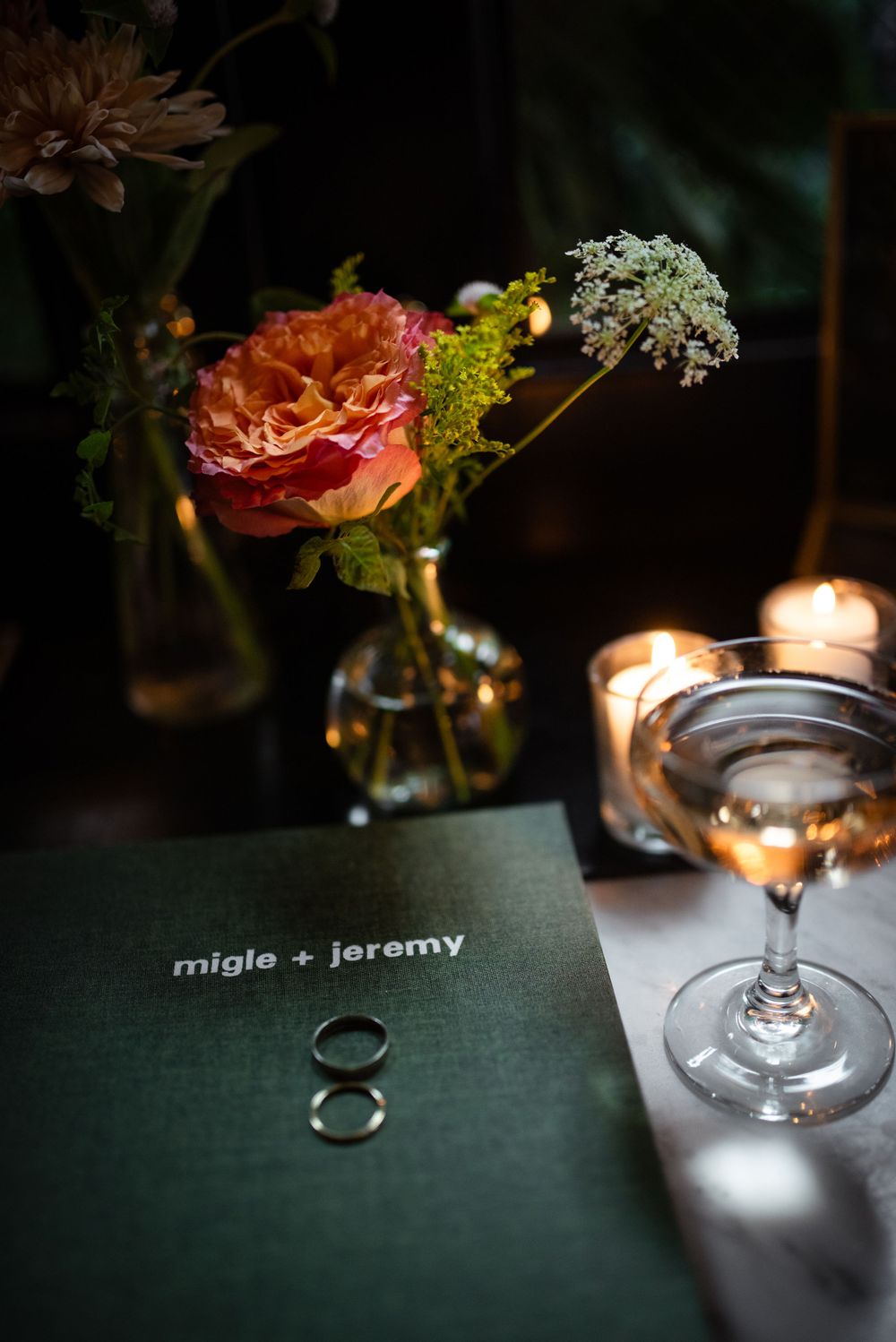 Wedding rings and floral centerpiece on a dark table with candlelight and a name card reading 'migle + jeremy'.