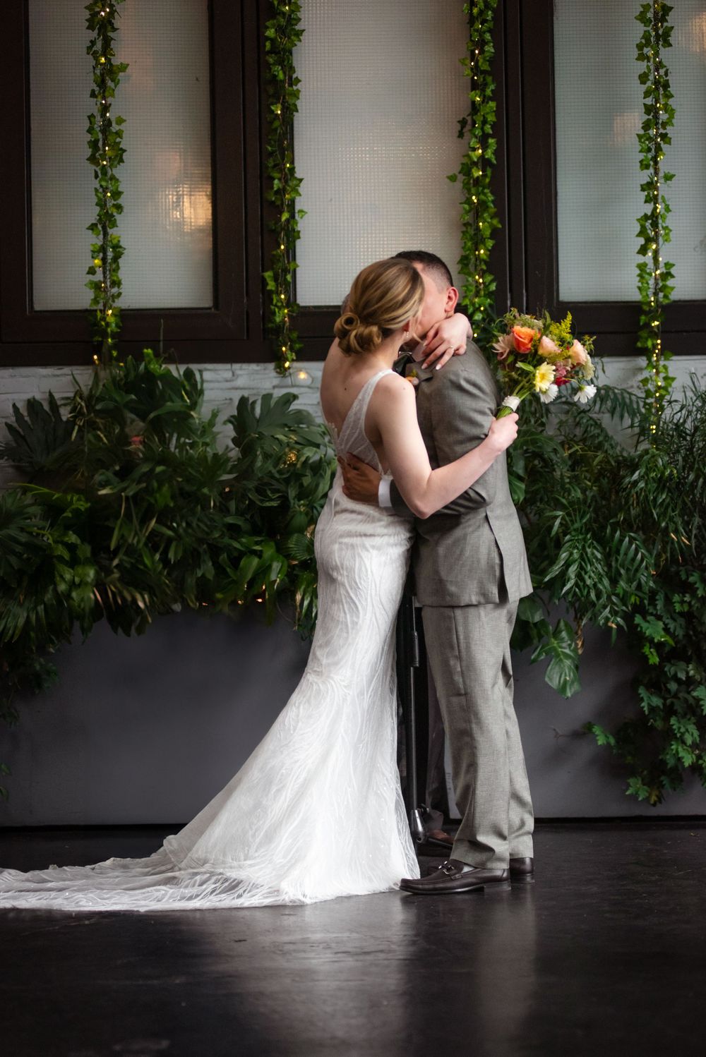 A couple embraces in formal wedding attire against a wall decorated with hanging greenery.