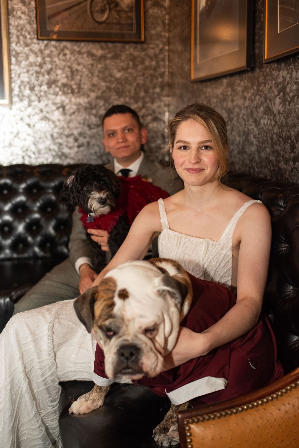 Two dogs sit with their owners on a leather couch in a cozy room with textured wallpaper.