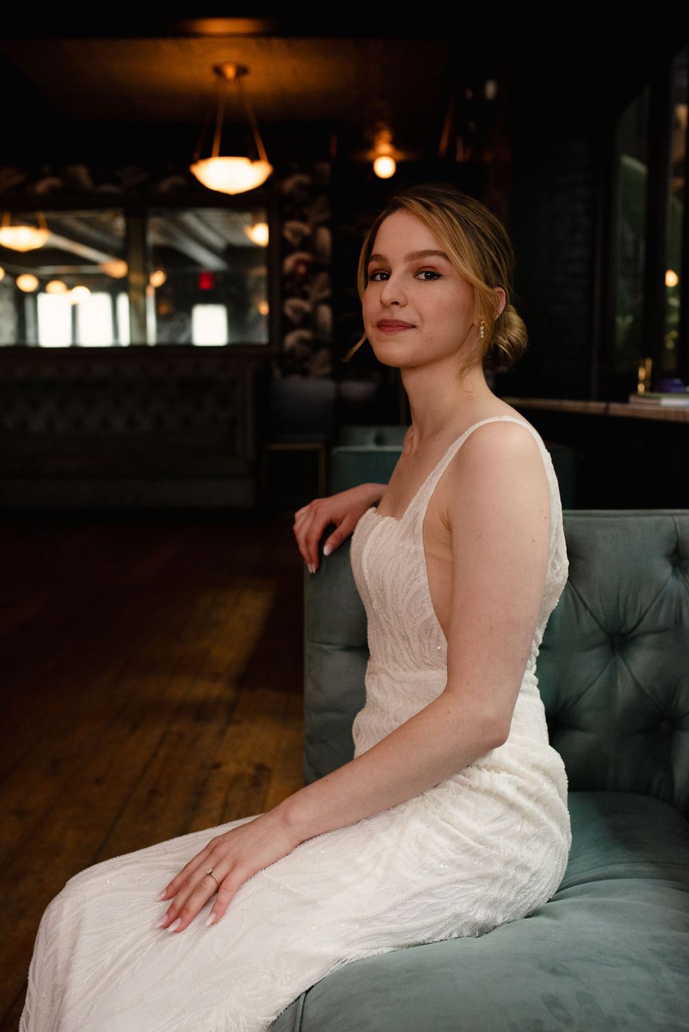 A person in a white lace wedding dress sits elegantly on a tufted green velvet couch in a dimly lit room.