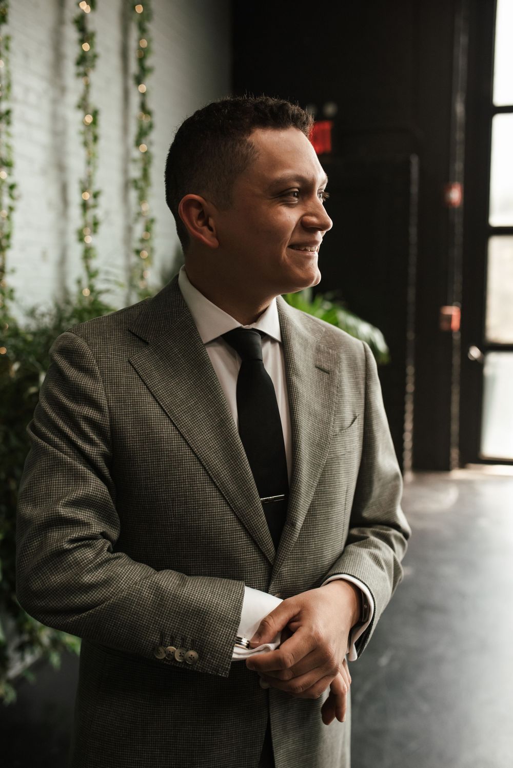 A person wearing a gray suit and tie smiles while standing in an elegant indoor setting with vine decorations.