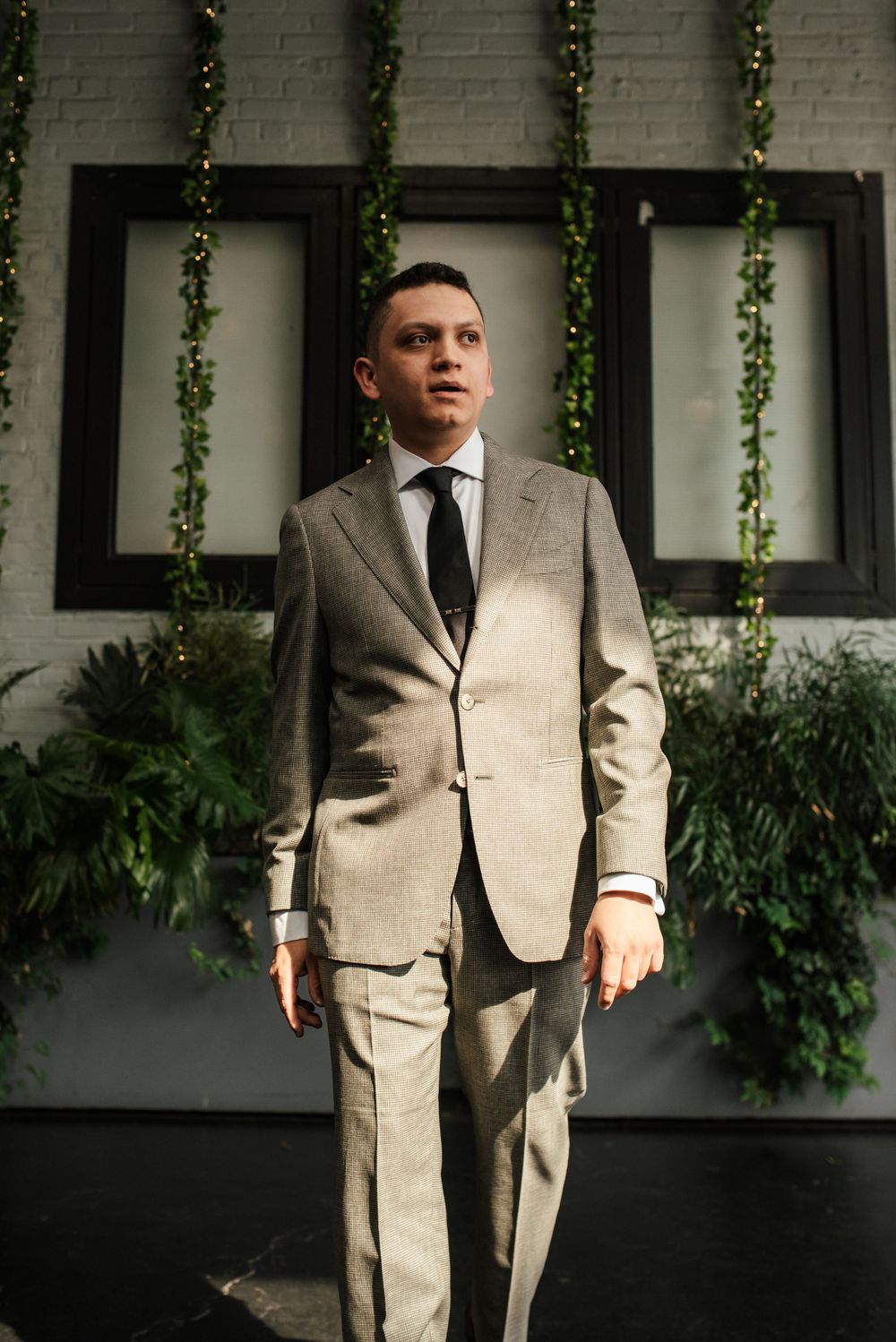 A person in a light gray suit stands confidently against a white wall decorated with hanging vines.