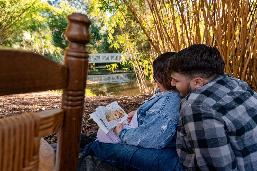 Couple sitting together against bamboo plants in a garden, reading a book outdoors near a pond.