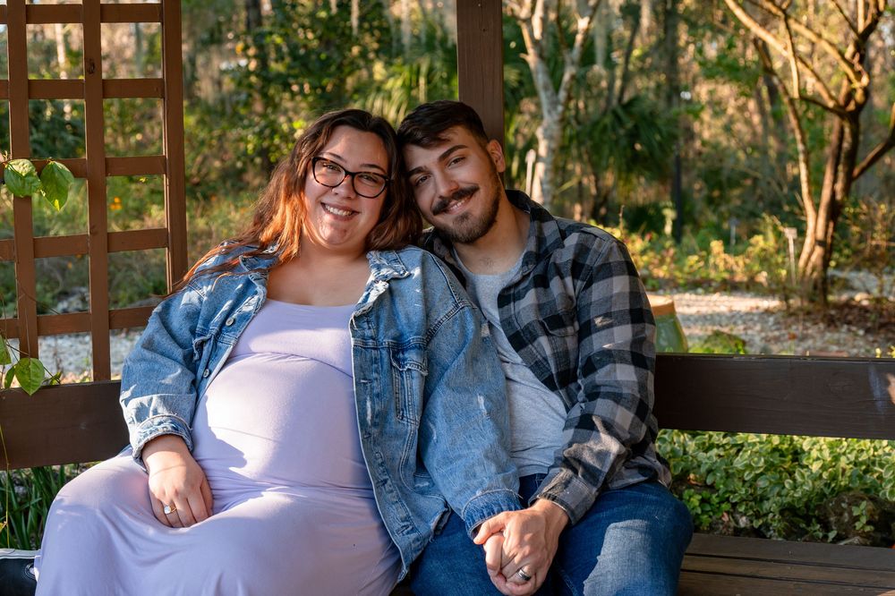 Smiling pregnant couple sitting together on a bench in a sunlit garden during a maternity photo shoot.