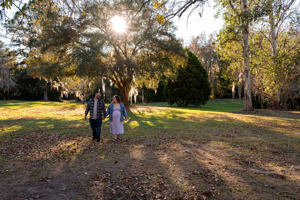 Couple holding hands walking through sunlit park with Spanish moss-draped oak trees during golden hour.