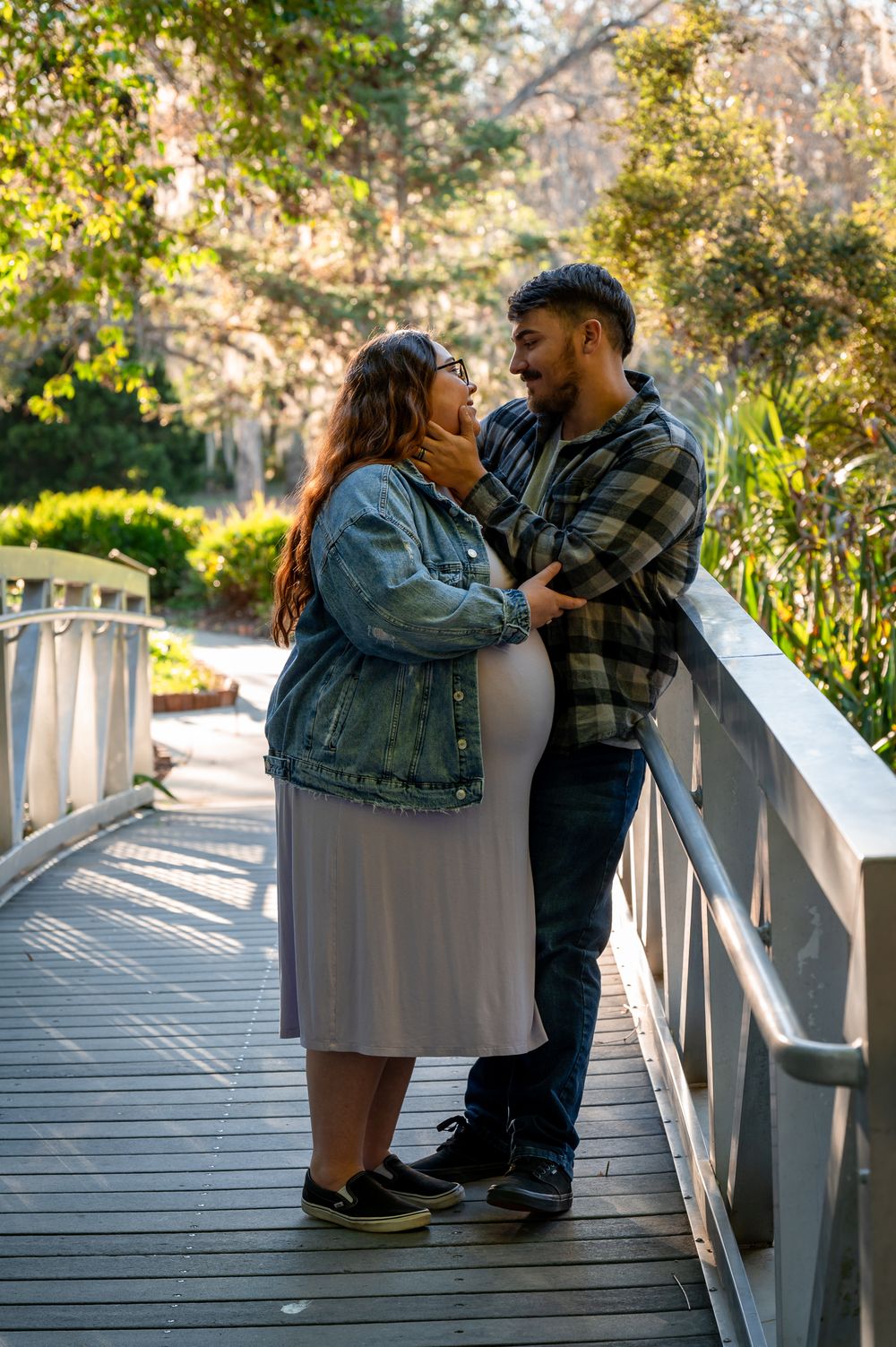 Pregnant couple sharing a tender moment on a wooden bridge surrounded by lush greenery in a park.