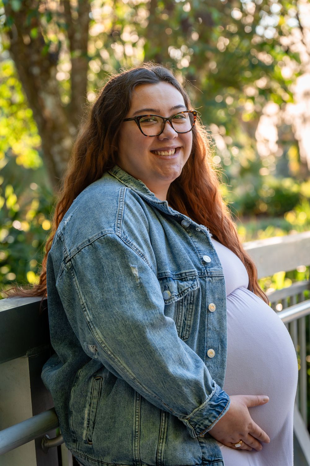 Smiling pregnant woman with red hair and glasses wearing a denim jacket outdoors on a bridge surrounded by greenery.
