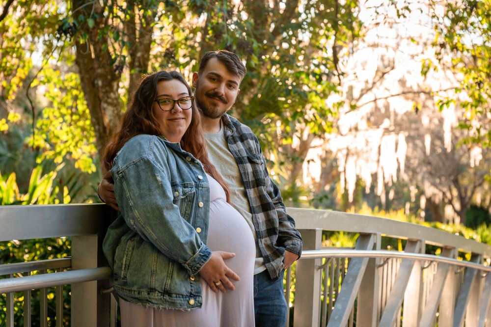 Pregnant couple posing on a wooden bridge surrounded by lush green trees during golden hour outdoors.