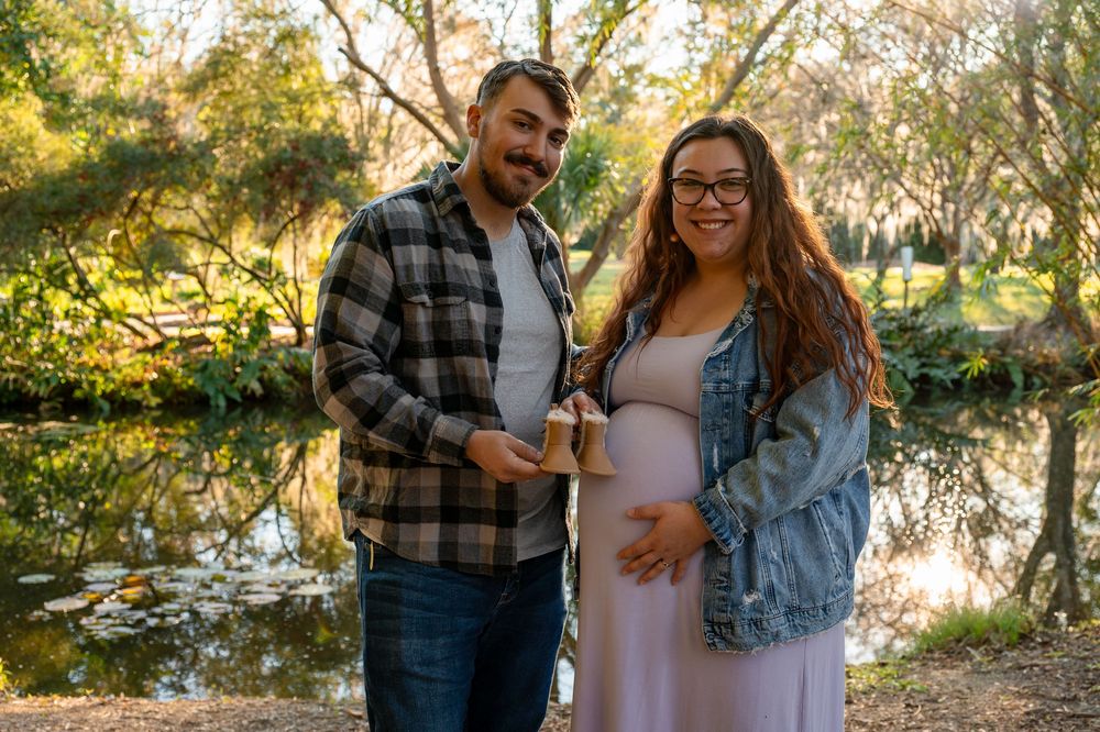Pregnant couple smiling together by a serene pond surrounded by lush trees in soft natural light.