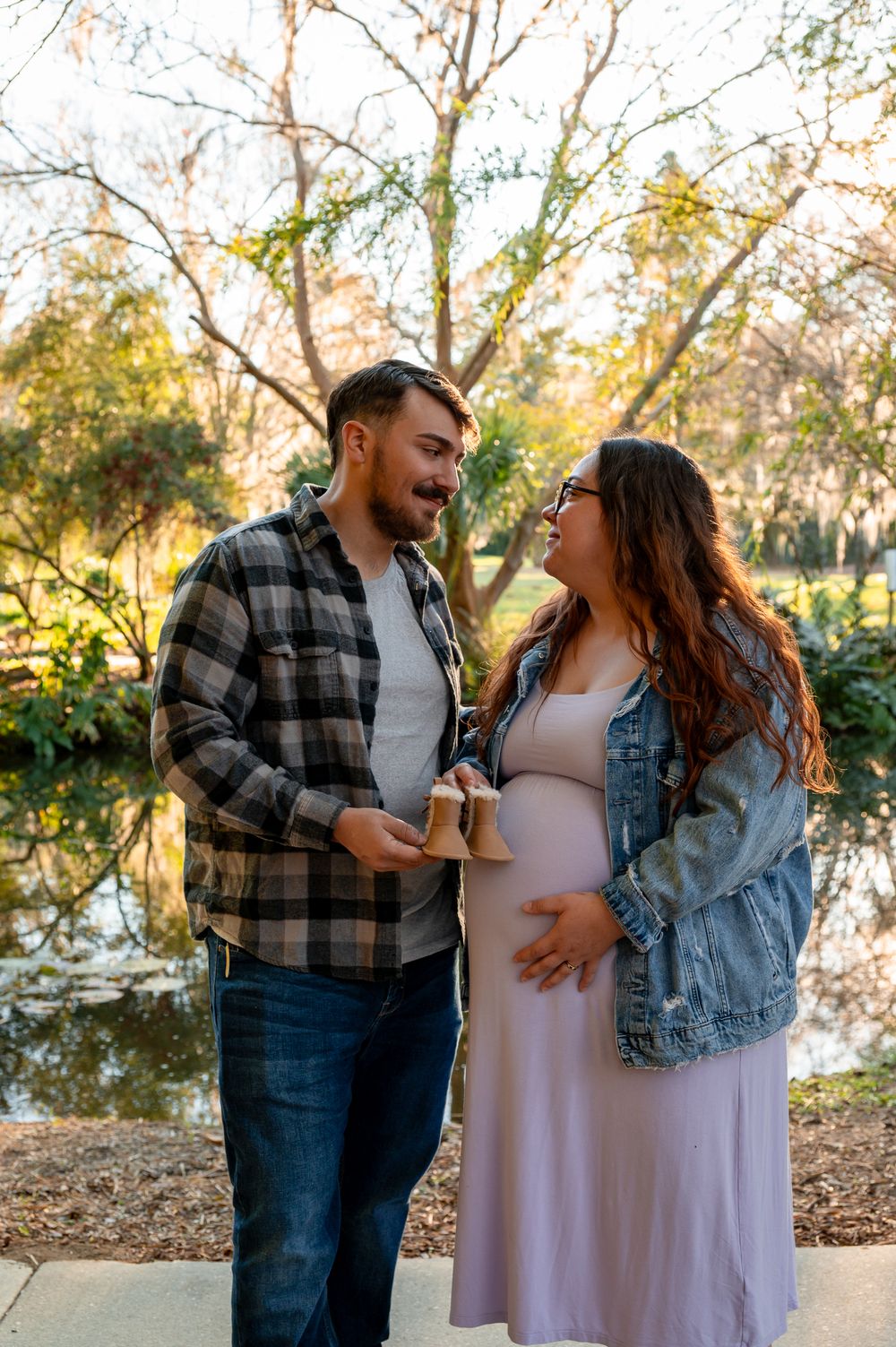 Expecting couple smiles at each other outdoors, man holds baby shoes as pregnant woman cradles her belly near a serene pond.