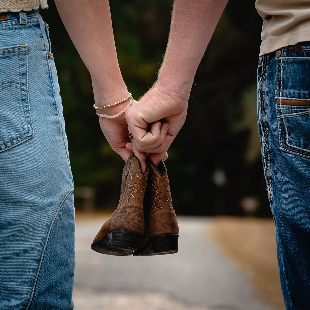 A close-up view of hands holding brown leather boots against a blurred outdoor background.