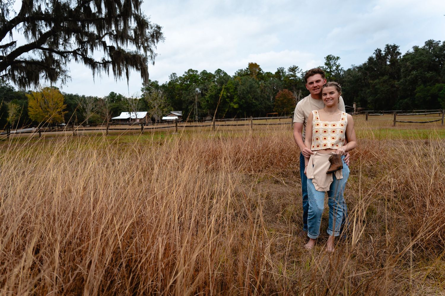 A couple stands together in a golden brown field of tall grass on an autumn day with trees in the background.