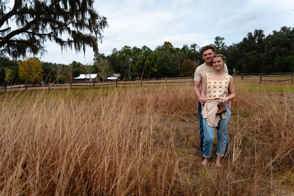 A couple stands together in a golden brown field of tall grass on an autumn day with trees in the background.