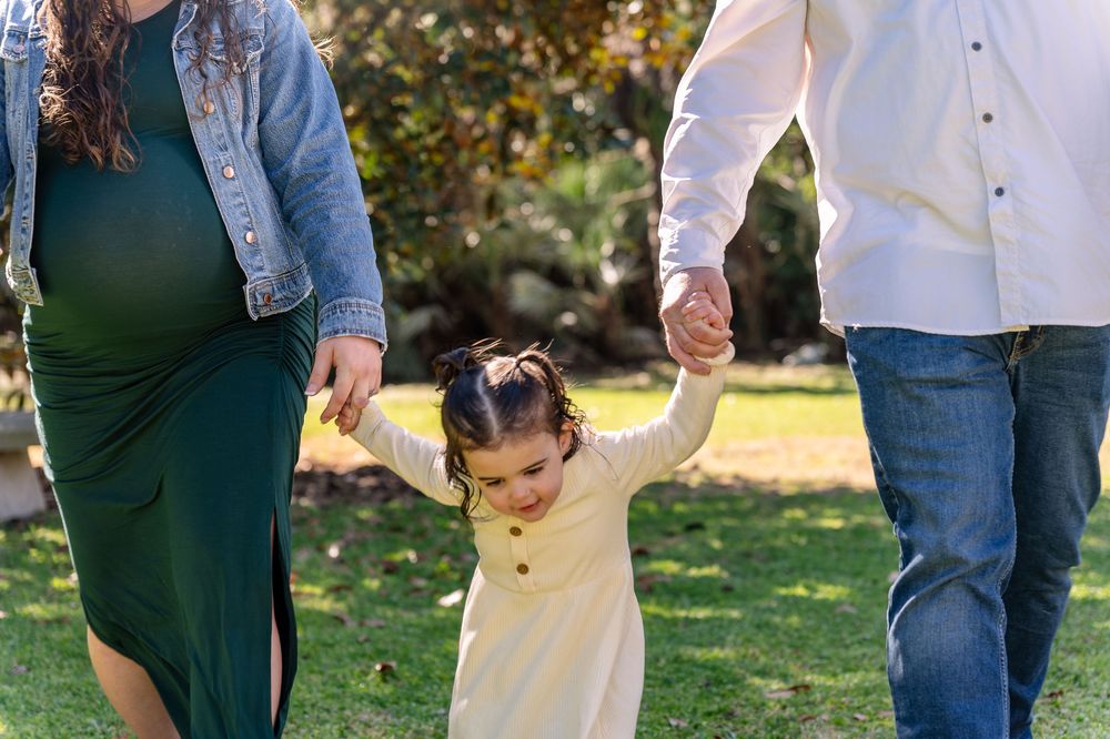 Pregnant woman and man walk holding hands with young toddler girl in green park setting.