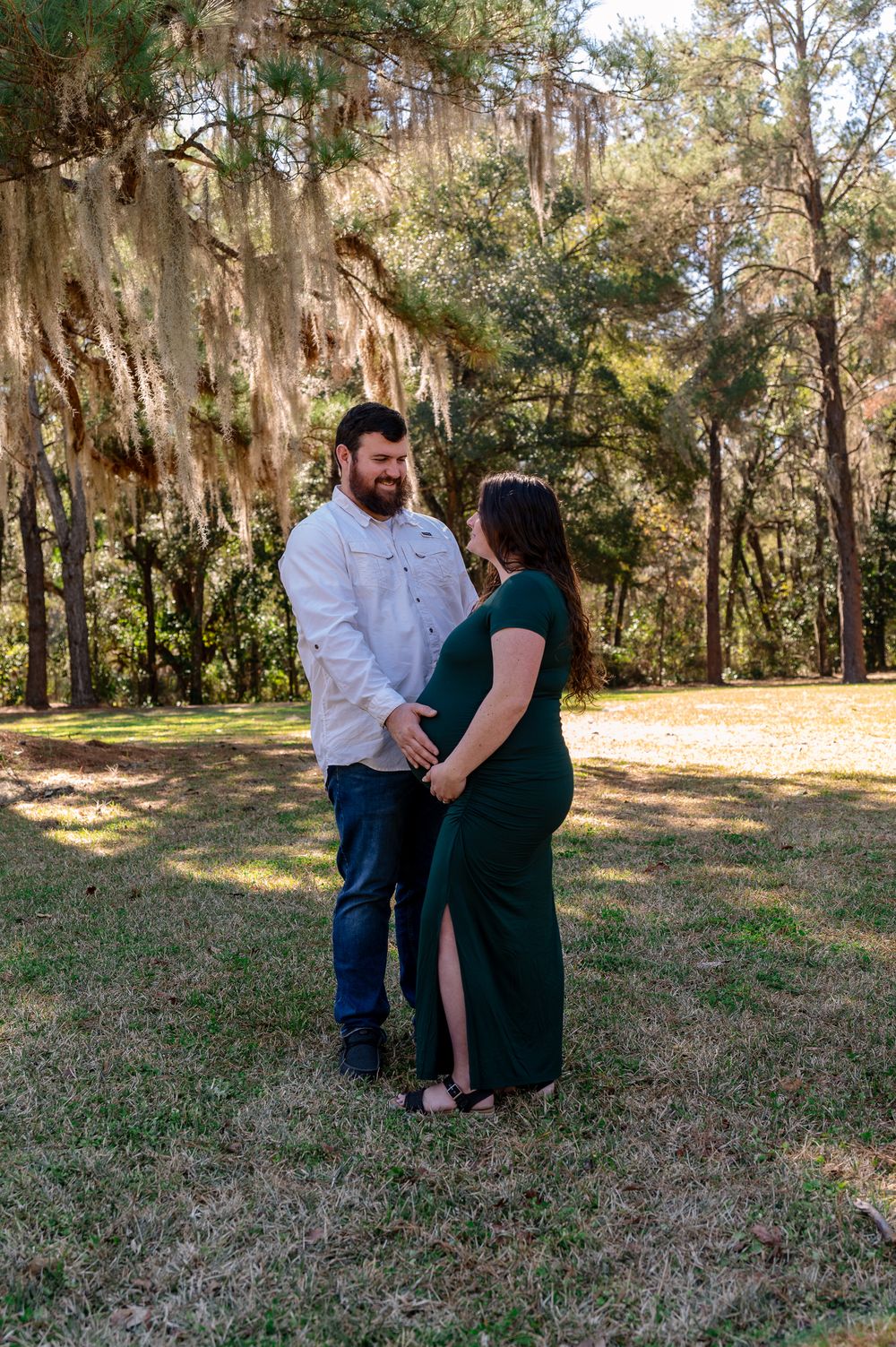 Couple holding hands in a sunny forest park, woman in green maternity dress, man in white shirt and jeans.