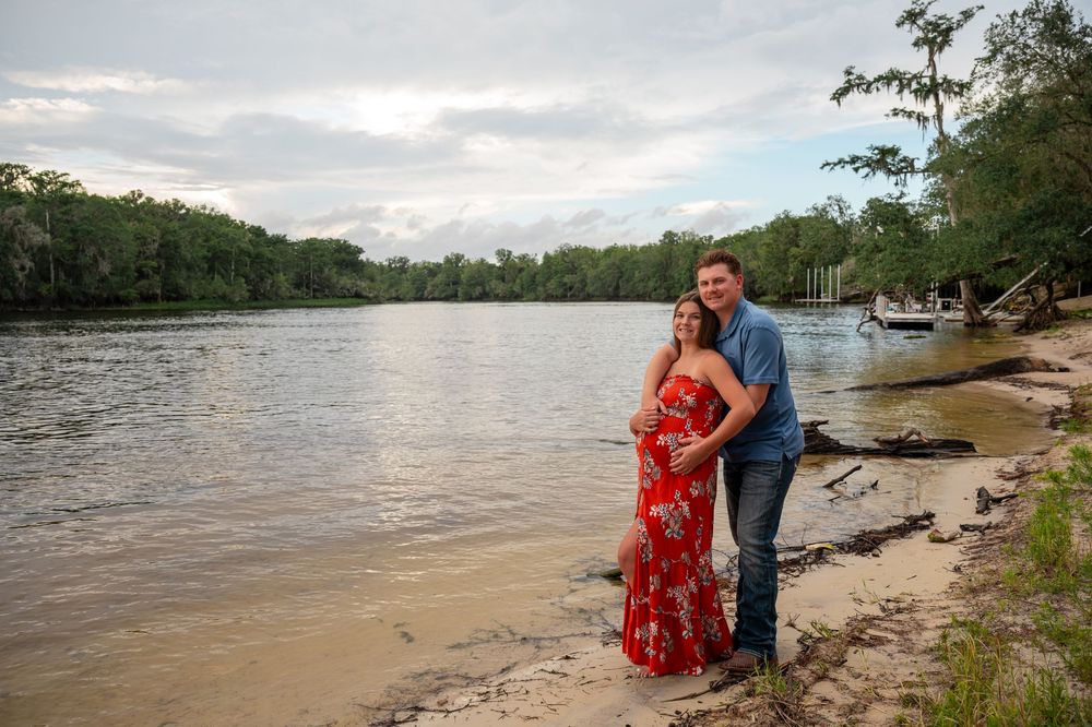 Pregnant couple posing on sandy riverbank at sunset, woman in red floral dress, man in blue shirt, lush trees behind.