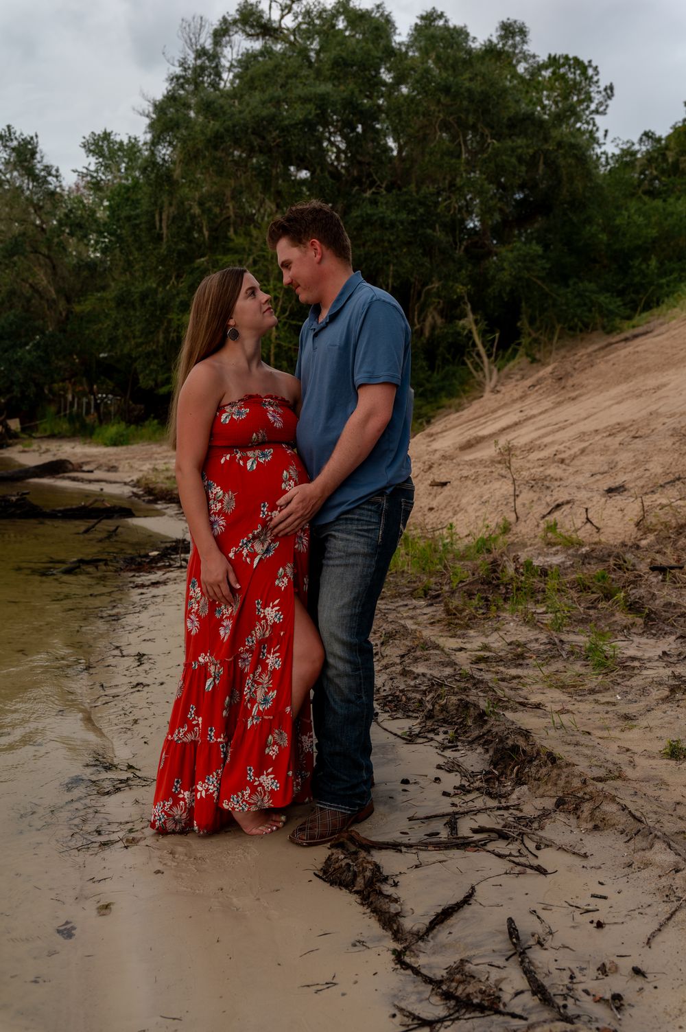 Couple sharing a romantic moment on a sandy riverbank, woman in red floral maxi dress, man in blue shirt and jeans.