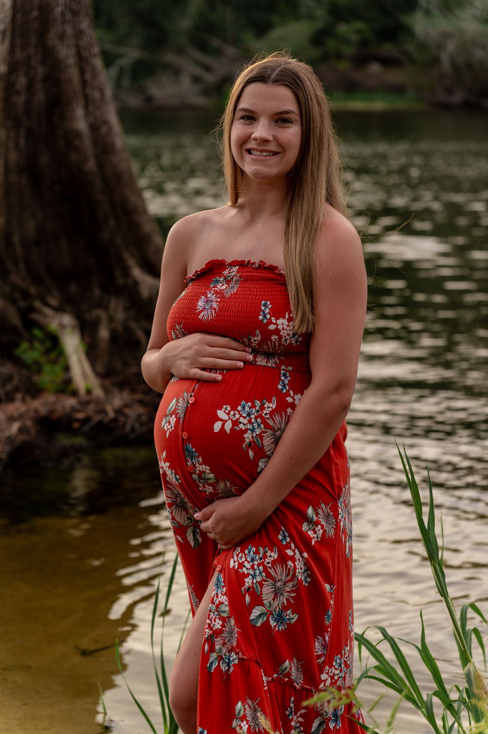 Smiling pregnant woman in red floral strapless maxi dress cradling baby bump outdoors near water.