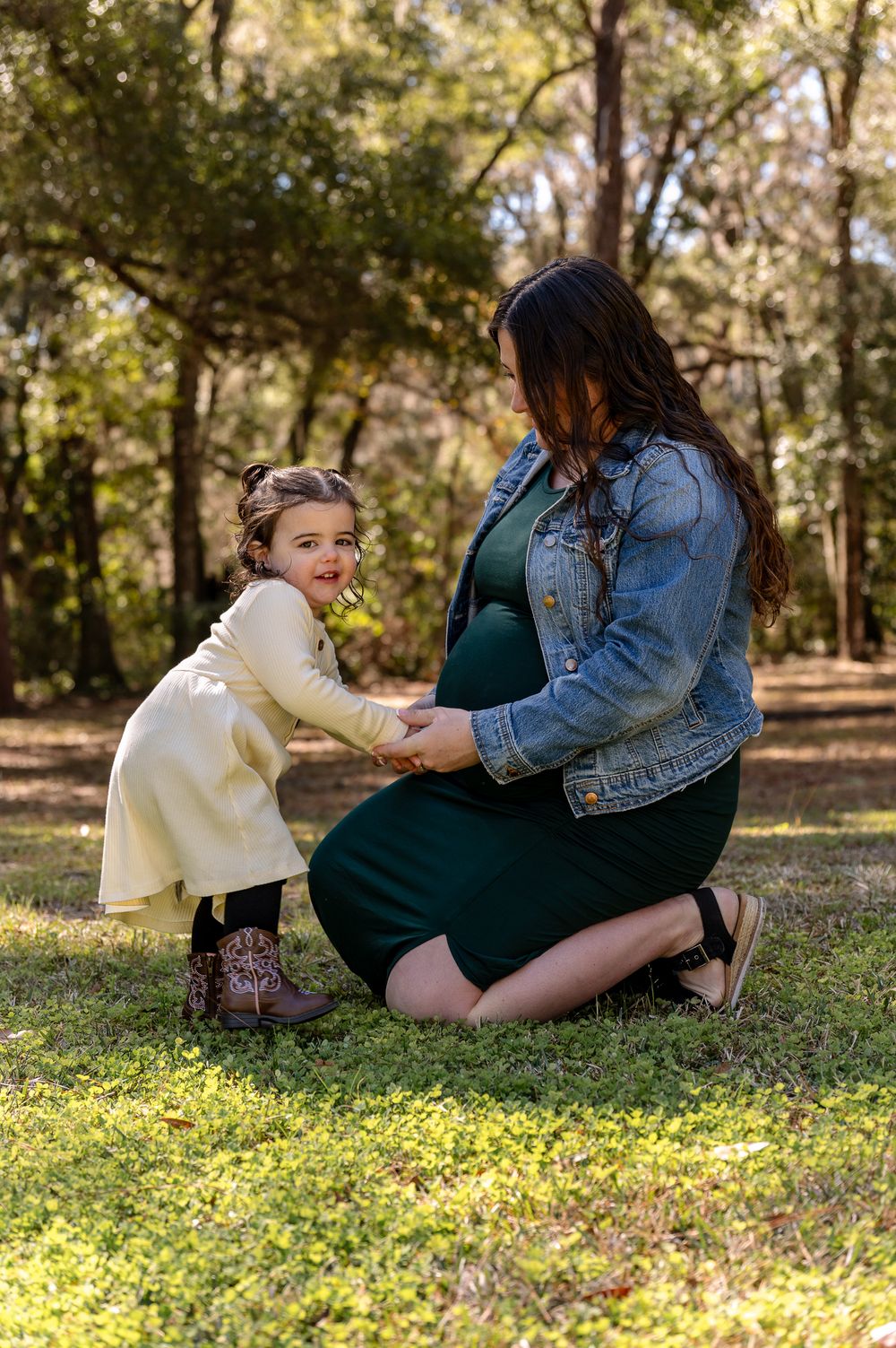 Pregnant woman kneeling outdoors with smiling toddler girl in cream coat and cowboy boots holding hands in wooded park.