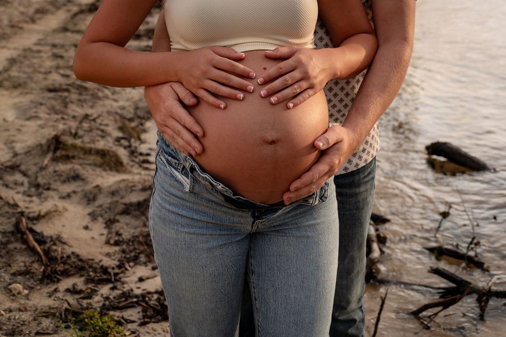 Pregnant woman in jeans with partner embracing her baby bump outdoors near sandy beach at sunset.