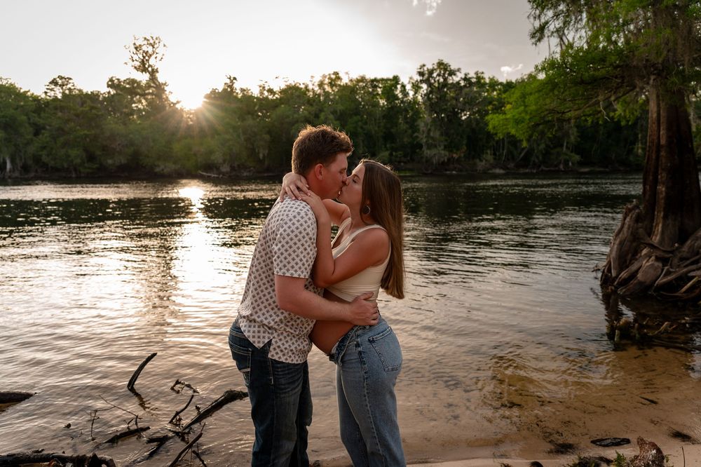 Couple sharing a romantic kiss by a sunlit river at golden hour, surrounded by lush green trees.