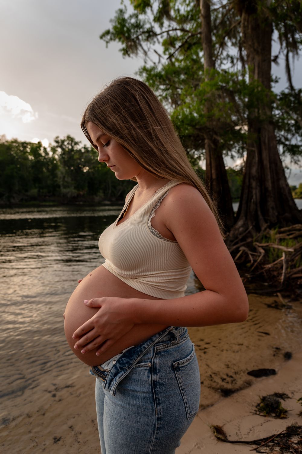 Pregnant woman in crop top and jeans cradling baby bump by a serene river at sunset.
