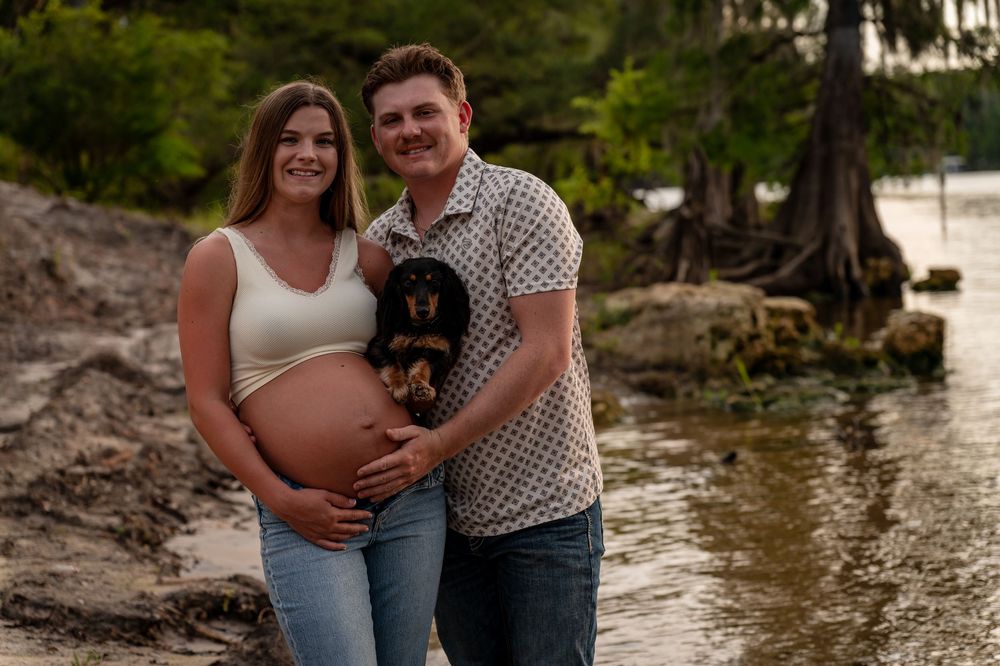 Pregnant couple smiling on a lakeside beach holding a small dog, surrounded by lush trees and calm water.