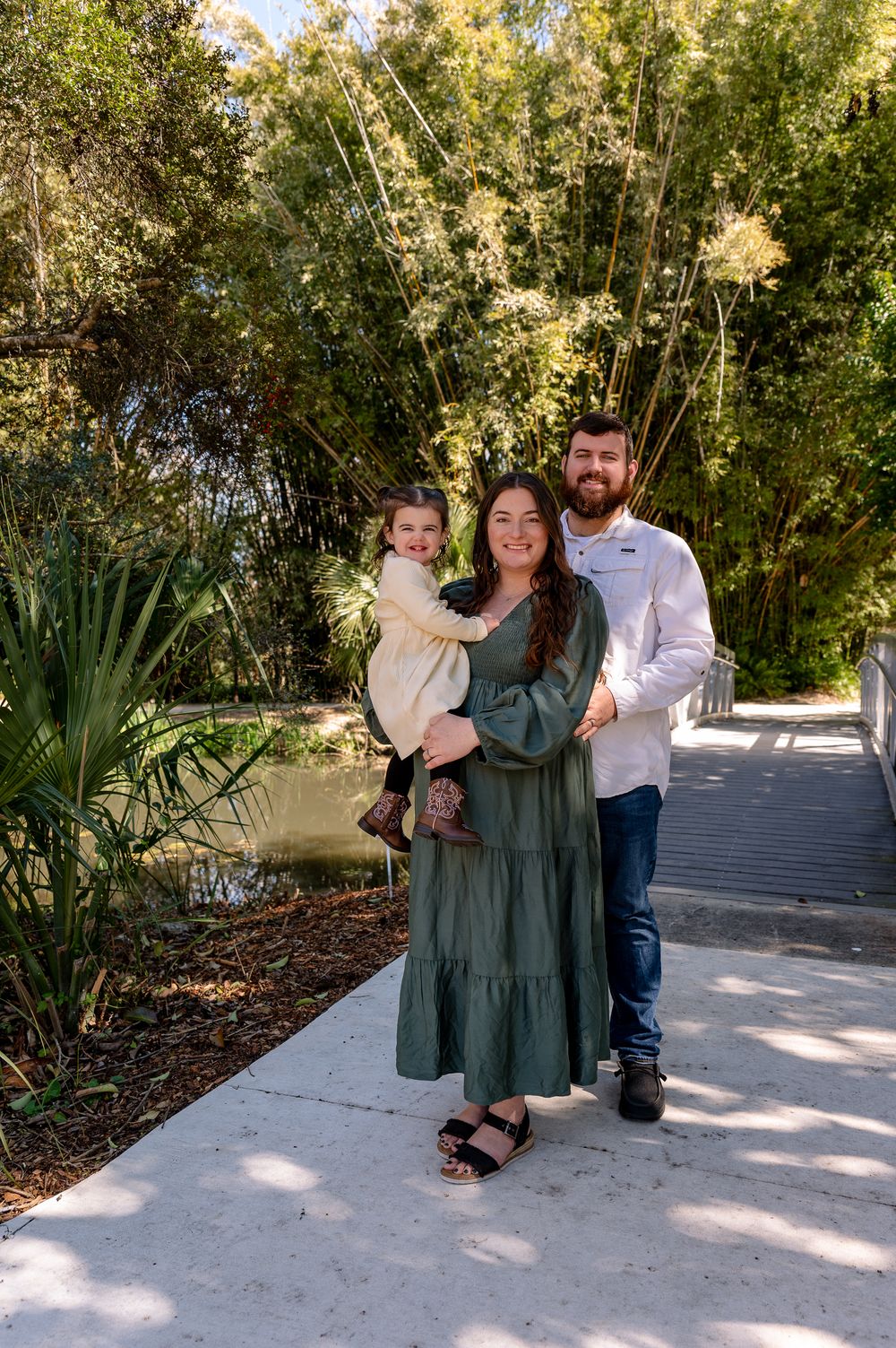Family of three posing on a garden path, mom in green dress holding toddler, dad in white shirt, lush trees behind.