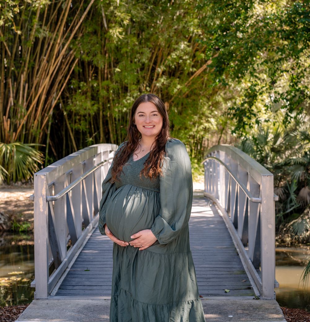 Smiling pregnant woman in green maxi dress poses on wooden bridge surrounded by lush bamboo trees.